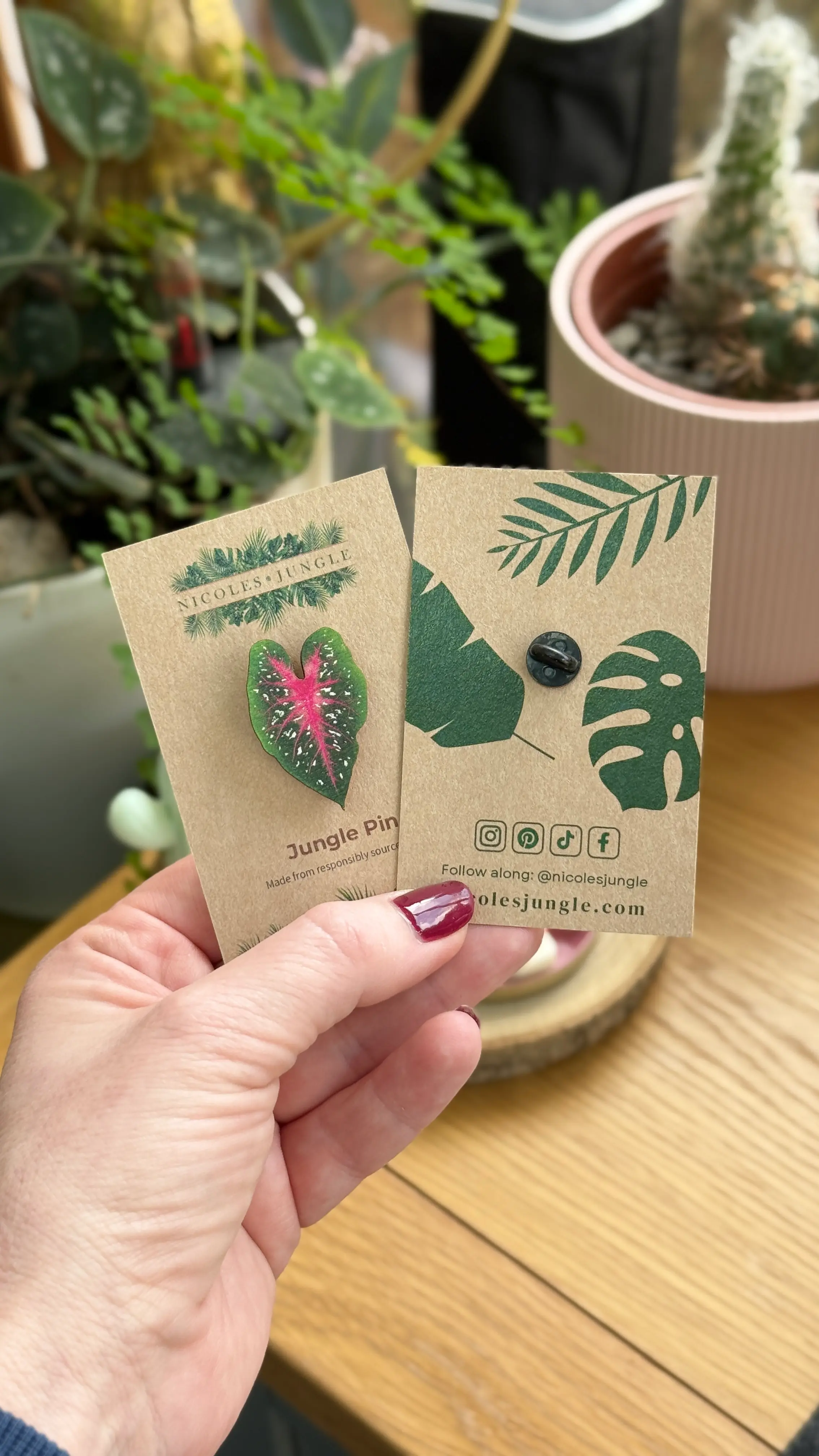 Front and back of a Nicole's Jungle wooden Caladium leaf pin held in a hand, shown on branded backing card.