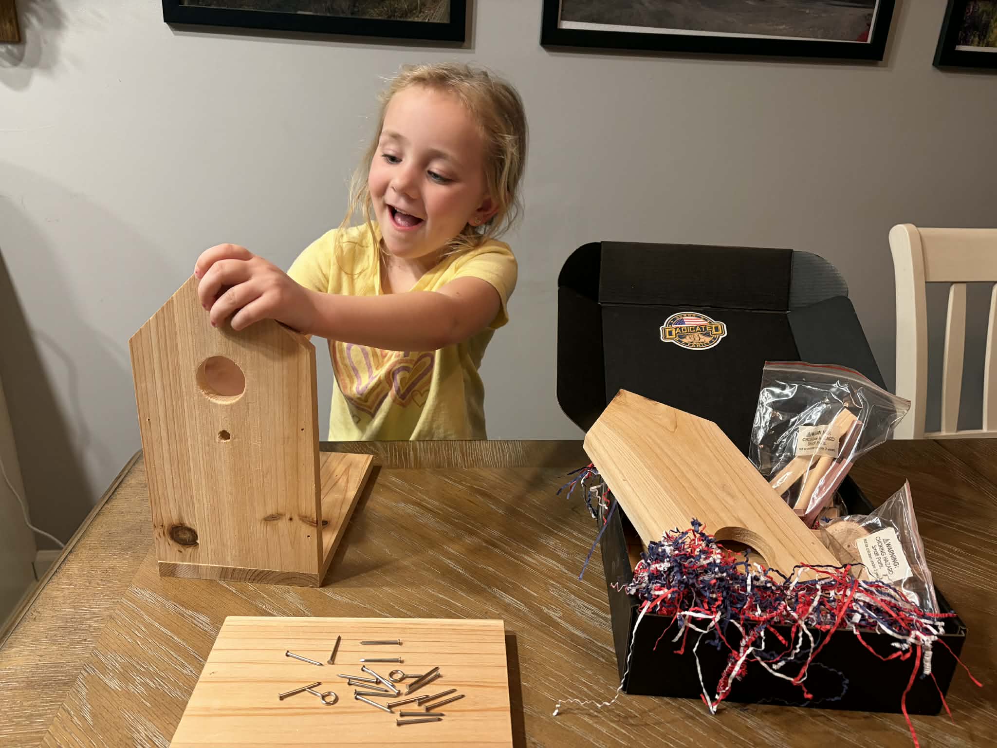 Young girl excitedly building a wooden birdhouse from her DADicated box with overflowing project materials on the table